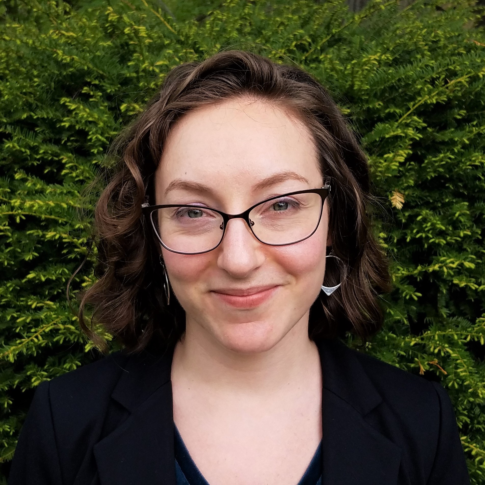 A close-up photo of a woman wearing a black blazer smiling and standing in front of green foliage.