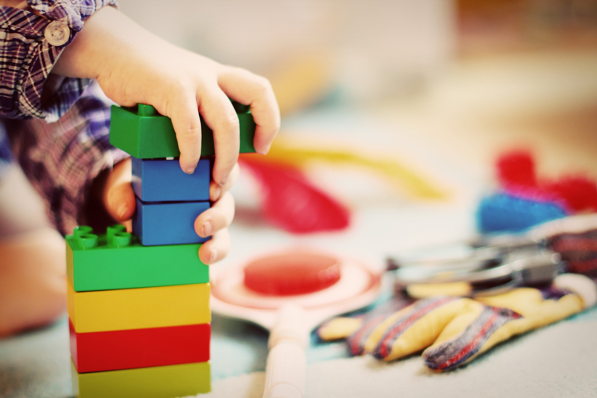 A close up photo of a child's hands building a tower out of Duplo style blocks. Other toys appear in the background.